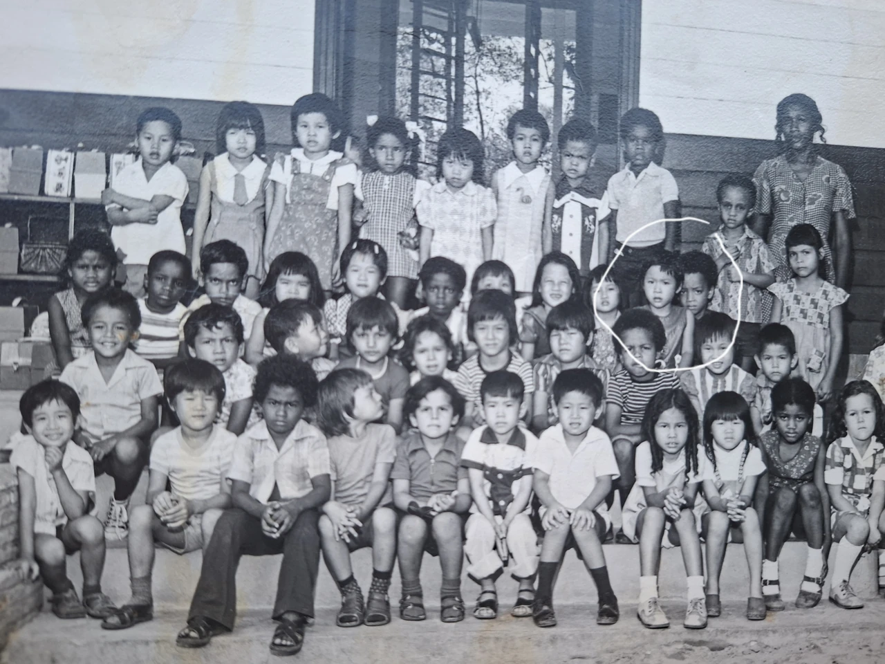 Historische zwart-wit groepsfoto van kinderen en een volwassene voor een houten gebouw.